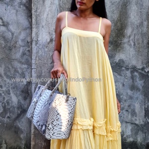 May include: A woman wearing a yellow sleeveless dress with a ruffled hem, holding a grey and white snakeskin handbag. The dress is made of a lightweight fabric and has a relaxed fit. The handbag has a top handle and a zip closure.