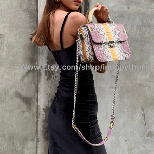 May include: A small, rectangular handbag with a snakeskin pattern in shades of pink, yellow, and white. The bag has a top handle and a chain strap. The bag is made of genuine python leather.