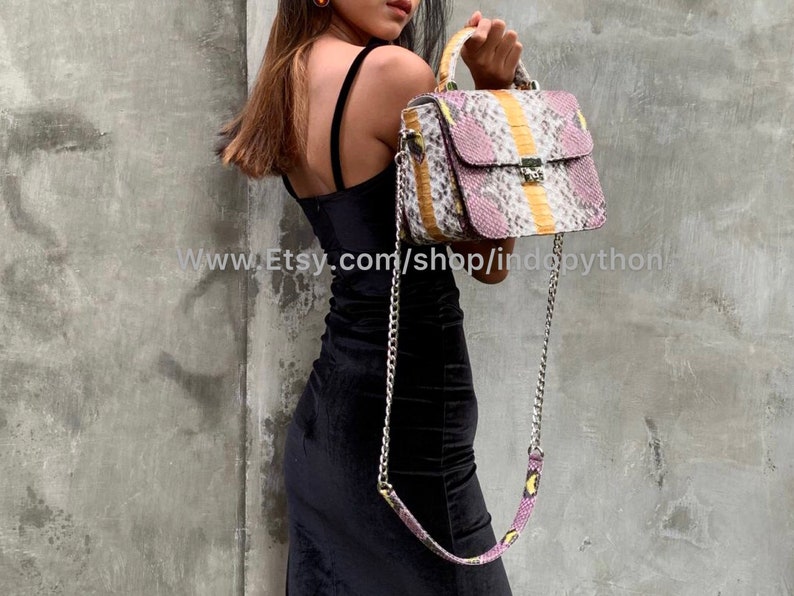 May include: A small, rectangular handbag with a snakeskin pattern in shades of pink, yellow, and white. The bag has a top handle and a chain strap. The bag is made of genuine python leather.