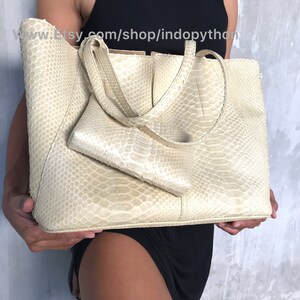May include: A large, off-white tote bag made of python skin. The bag has a top zipper closure and two top handles. The bag is being held by a person.