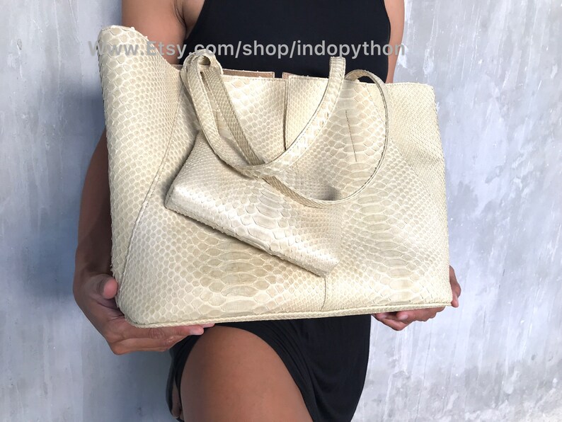 May include: A large, off-white tote bag made of python skin. The bag has a top zipper closure and two top handles. The bag is being held by a person.