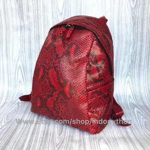Könnte beinhalten: Ein roter Schlangenleder-Rucksack mit schwarzen Akzenten. Der Rucksack hat einen Reißverschluss und verstellbare Träger.