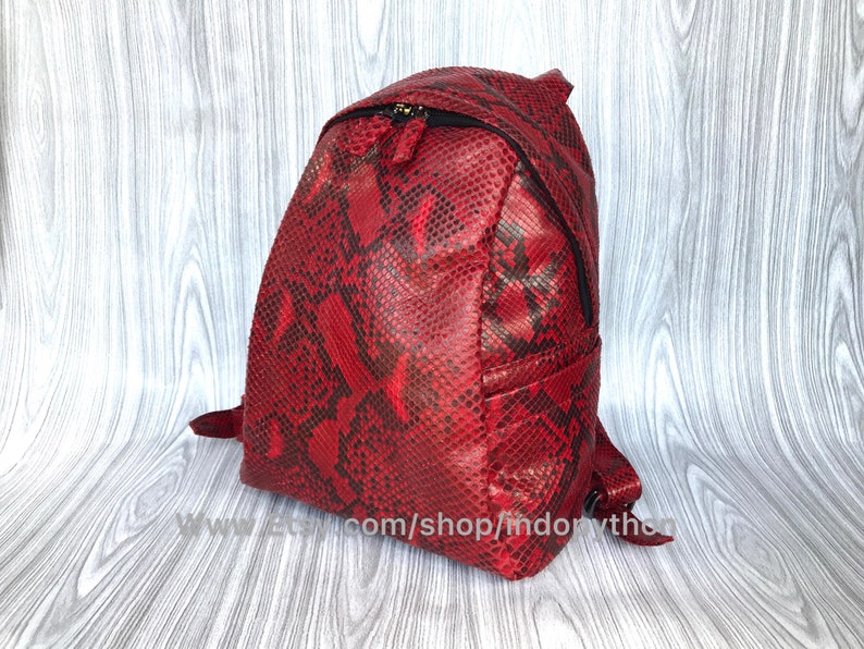 May include: A red snakeskin backpack with black accents. The backpack has a zipper closure and adjustable straps.