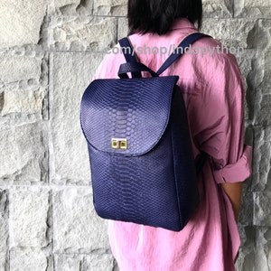 May include: A dark blue snakeskin backpack with a flap closure and gold hardware. The backpack is being worn by a person.
