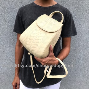 May include: A small, off-white leather backpack with a flap closure and a top handle. The backpack is made of python skin and has a textured surface.