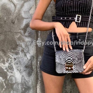 May include: A black and white snakeskin crossbody bag with a silver chain strap and a silver clasp. The bag is being worn by a person wearing a black and white striped top and black denim shorts.