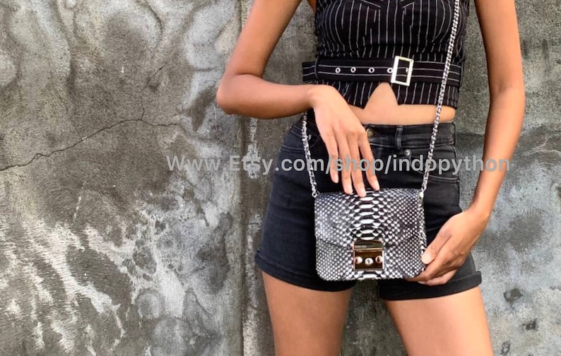 May include: A black and white snakeskin crossbody bag with a silver chain strap and a silver clasp. The bag is being worn by a person wearing a black and white striped top and black denim shorts.