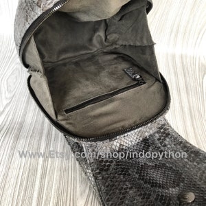 K&ouml;nnte beinhalten: Eine schwarze und graue Schlangenlederhandtasche mit Rei&szlig;verschluss und einer kleinen Innentasche. Die Tasche ist offen und das Innere ist mit grauem Stoff ausgekleidet.