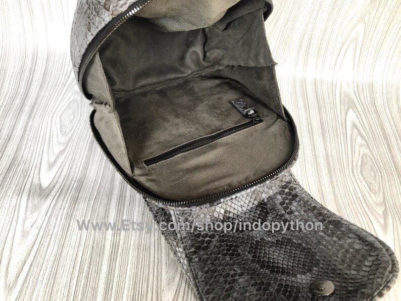 K&ouml;nnte beinhalten: Eine schwarze und graue Schlangenlederhandtasche mit Rei&szlig;verschluss und einer kleinen Innentasche. Die Tasche ist offen und das Innere ist mit grauem Stoff ausgekleidet.