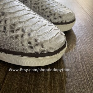 K&ouml;nnte beinhalten: Nahaufnahme eines Paares wei&szlig;er Slipper aus Pythonleder mit braunem Besatz. Die Schuhe haben eine wei&szlig;e Gummisohle und ein strukturiertes Muster. Der Text "www.Etsy.com/shop/indopython" ist am unteren Rand des Bildes sichtbar.