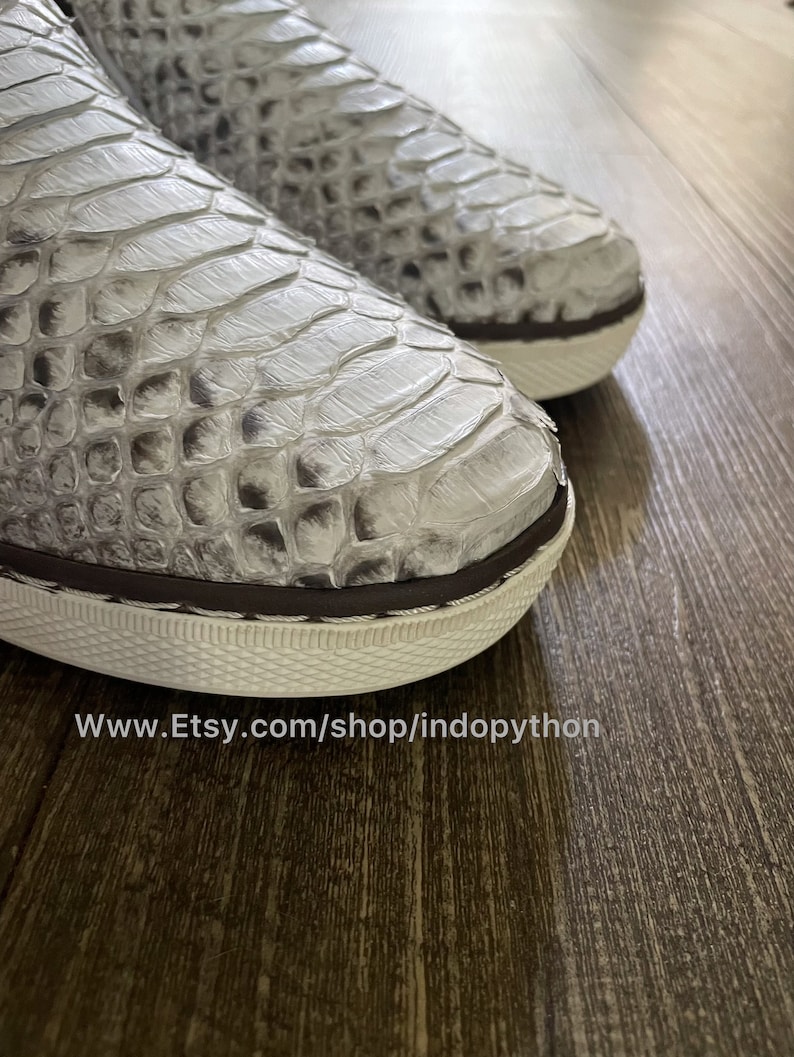 K&ouml;nnte beinhalten: Nahaufnahme eines Paares wei&szlig;er Slipper aus Pythonleder mit braunem Besatz. Die Schuhe haben eine wei&szlig;e Gummisohle und ein strukturiertes Muster. Der Text "www.Etsy.com/shop/indopython" ist am unteren Rand des Bildes sichtbar.