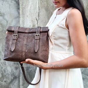 Könnte beinhalten: Eine braune Ledertasche mit einem Klappverschluss und zwei Schnallen. Die Tasche ist aus Pythonleder und hat einen langen Riemen.