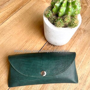 Op de afbeelding: Een donkergroene, getextureerde leren brillenkoker met een zilveren drukknoopsluiting. De koker is rechthoekig met een gebogen klep. Een kleine cactus in een witte pot staat op de achtergrond.
