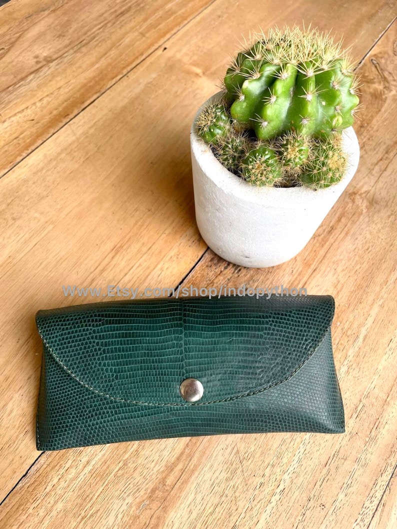 Op de afbeelding: Een donkergroene, getextureerde leren brillenkoker met een zilveren drukknoopsluiting. De koker is rechthoekig met een gebogen klep. Een kleine cactus in een witte pot staat op de achtergrond.