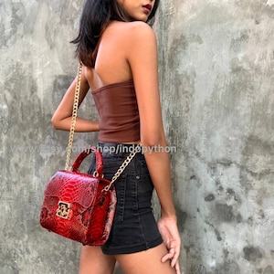 Könnte beinhalten: Eine rote Schlangenleder-Handtasche mit einer goldenen Kettenriemen und einem goldenen Verschluss. Die Tasche hat einen oberen Griff und wird von einer Person getragen.