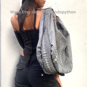 Può includere: Una borsa a mano in pelle di serpente grigia con riflessi argentati. La borsa è indossata sulla spalla di una persona che indossa una maglietta nera e jeans neri.