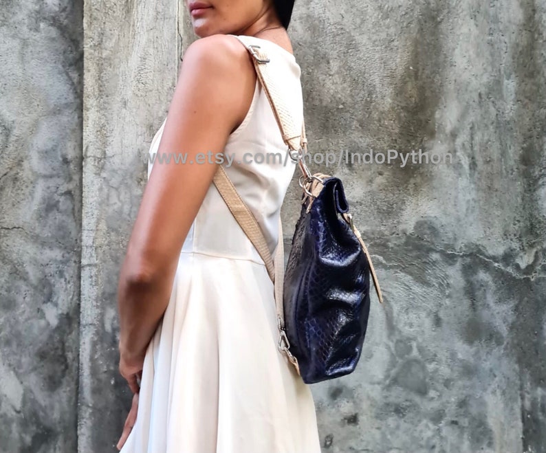 May include: A navy blue leather backpack with a snakeskin pattern. The backpack has a beige adjustable strap and a zipper closure.