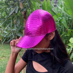 Può includere: Un berretto da baseball rosa in pelle di serpente con una finitura lucida. Il berretto è indossato da una persona con lunghi capelli scuri. Il testo "www.Etsy.com/shop/indopython" è visibile sul visore del berretto.