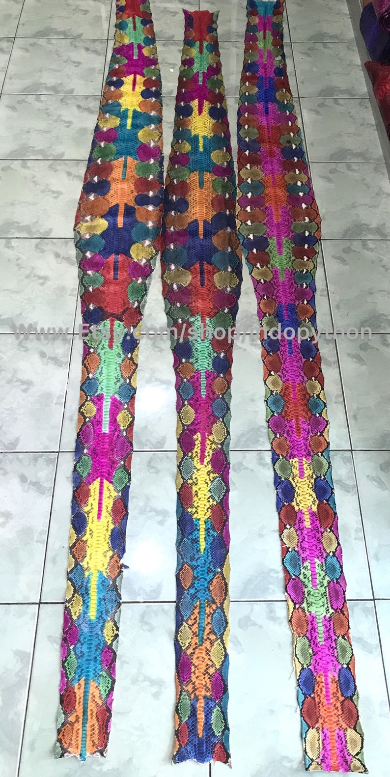 May include: Three long strips of colorful snakeskin, each with a different pattern. The skins are arranged vertically and are all about the same length. The colors are bright and vibrant, including red, orange, yellow, green, blue, and purple.