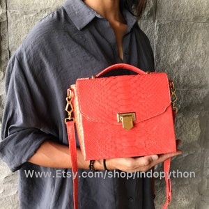 May include: A bright red satchel handbag made of snakeskin with a gold clasp and a long adjustable strap. The bag is held in the hands of a person wearing a dark grey shirt.