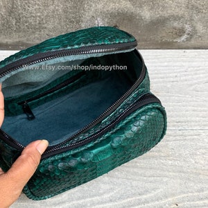 May include: A green snakeskin makeup bag with a black zipper. The bag is open and the inside is lined with a dark green fabric. The bag is sitting on a light brown surface.