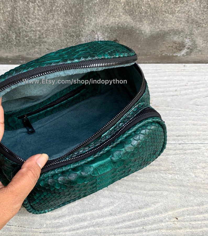 May include: A green snakeskin makeup bag with a black zipper. The bag is open and the inside is lined with a dark green fabric. The bag is sitting on a light brown surface.