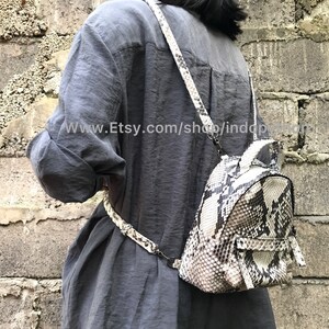 K&ouml;nnte beinhalten: Ein kleiner, wei&szlig;er und schwarzer Schlangenleder-Rucksack mit Rei&szlig;verschluss. Der Rucksack wird von einer Person getragen, die ein graues Hemd tr&auml;gt.