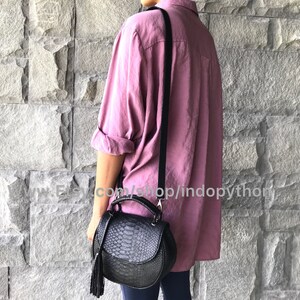 May include: A black leather satchel bag with a snakeskin pattern and a tassel. The bag has a top handle and a long strap. The bag is being worn by a person wearing a pink shirt.