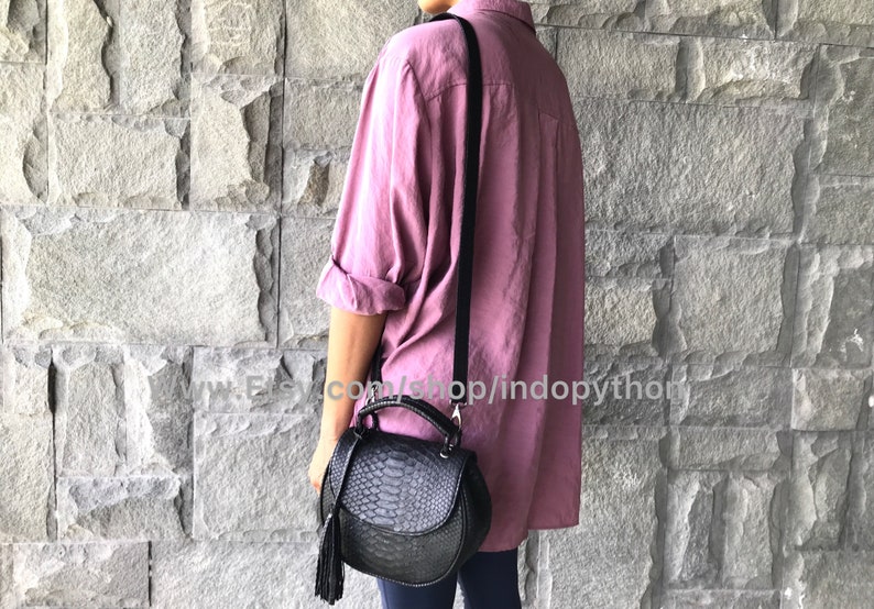 May include: A black leather satchel bag with a snakeskin pattern and a tassel. The bag has a top handle and a long strap. The bag is being worn by a person wearing a pink shirt.