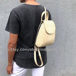 May include: A cream-colored leather backpack with a snakeskin pattern. The backpack has a flap closure and a zippered pocket on the front. The backpack is being worn by a person.
