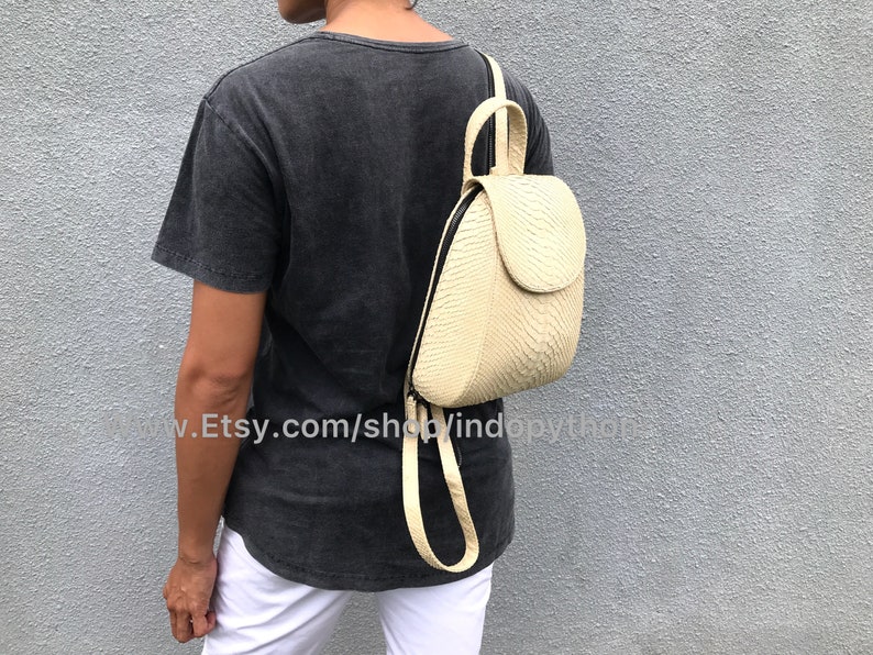 May include: A cream-colored leather backpack with a snakeskin pattern. The backpack has a flap closure and a zippered pocket on the front. The backpack is being worn by a person.