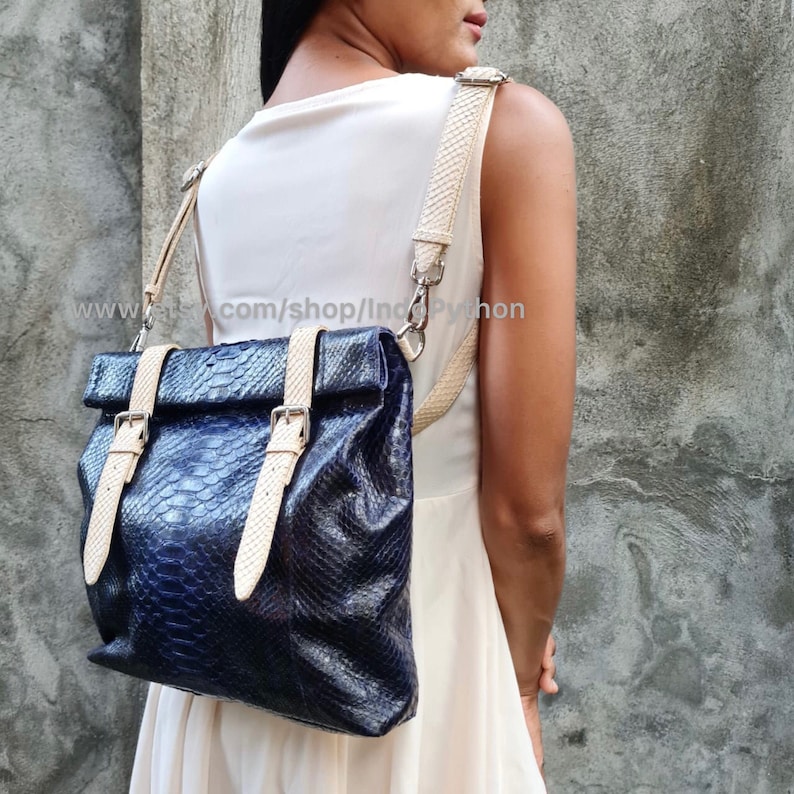 May include: A blue python leather backpack with cream-colored straps and buckles. The bag has a fold-over flap with a buckle closure.