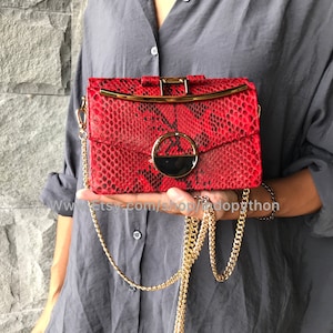 Puede incluir: Un bolso de mano de piel de serpiente roja con una correa de cadena dorada y un cierre de metal dorado. El bolso tiene un diseño circular en la parte delantera con un acento negro y dorado.