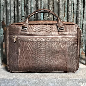 Pode incluir: Uma pasta de couro marrom com padrão de pele de cobra. A bolsa tem uma alça superior, um bolso frontal com zíper e ferragens prateadas. A pasta retangular parece ser feita de materiais de alta qualidade.