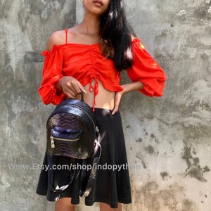 Pu&ograve; includere: Uno zaino nero e iridescente con un motivo a pelle di serpente strutturato. Lo zaino ha una piccola tasca frontale con cerniera dorata e spallacci regolabili. L'immagine mostra anche un top rosso con spalle scoperte e una gonna nera. L'URL del negozio Etsy &egrave; visibile.