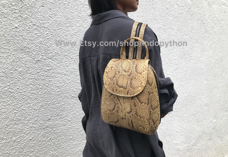 Puede incluir: Una peque&ntilde;a mochila de piel de serpiente de color beige con correas negras. La mochila tiene un cierre de solapa y un bolsillo con cremallera en la parte delantera. www.Etsy.com/shop/mdopython