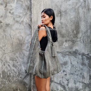 May include: A gray snakeskin handbag with a tassel. The bag is worn over the shoulder of a person wearing a black tank top.