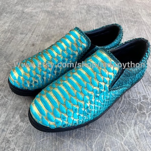 Peut inclure: Paire de chaussures à enfiler avec un motif peau de serpent turquoise et doré. Les chaussures ont une semelle noire et un bout arrondi. Le matériau supérieur présente un motif en forme d'écailles.