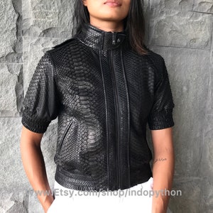 Puede incluir: Una chaqueta de cuero negra con cierre de cremallera y mangas cortas. La chaqueta está hecha de piel de pitón y tiene un patrón texturizado.