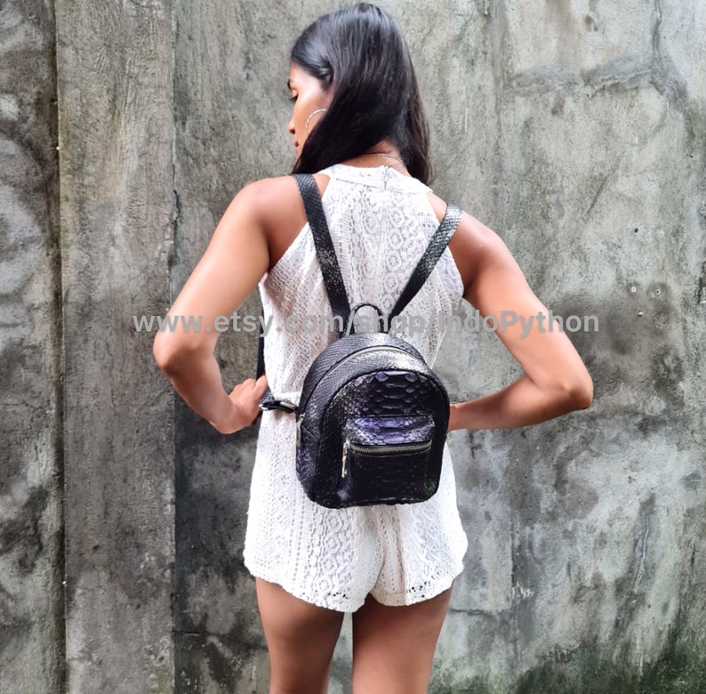 May include: A woman wearing a white dress with a black leather backpack with a snakeskin pattern. The backpack has a zipper closure and adjustable straps.