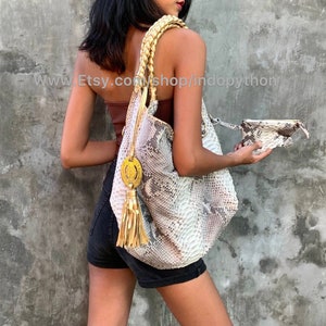 Può includere: Una borsa a mano in pelle di serpente bianca e marrone con un pendente dorato a forma di nappa e una tracolla intrecciata. La borsa è tenuta da una persona che indossa una canottiera marrone e pantaloncini neri.