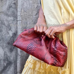 May include: A red python skin clutch purse with a gathered, pleated design. The purse is held in the hand of a person wearing a yellow dress.