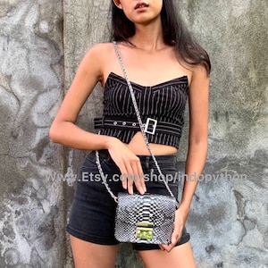 May include: A black and white snakeskin crossbody bag with a silver chain strap. The bag has a gold clasp and is being worn by a person wearing a black and white striped top and black shorts.