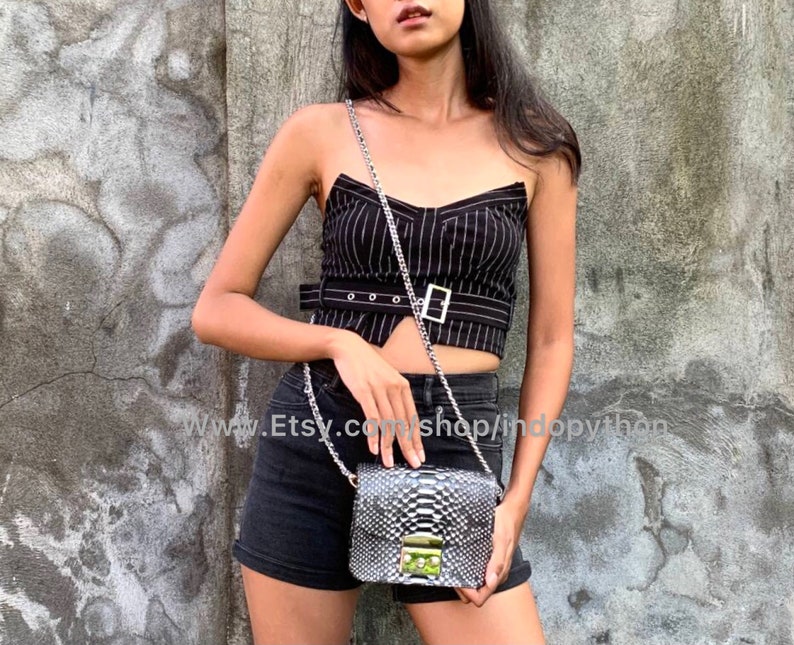 May include: A black and white snakeskin crossbody bag with a silver chain strap. The bag has a gold clasp and is being worn by a person wearing a black and white striped top and black shorts.