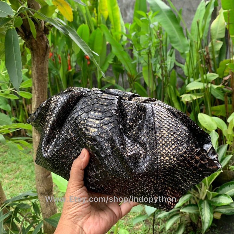 K&ouml;nnte beinhalten: Eine schwarze Clutch aus Pythonleder mit goldenen Akzenten. Die Clutch ist in der Mitte gefaltet und wird in einer Hand gehalten. Die Clutch hat eine strukturierte Oberfl&auml;che und einen goldenen Schimmer.