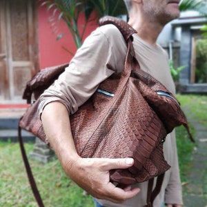 Puede incluir: Un bolso de cuero marrón con un patrón texturizado y escamoso. El bolso tiene una correa larga y un cierre de cremallera.