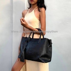 May include: A black leather tote bag with a snakeskin pattern and a tassel. The bag has two top handles and a zipper closure.