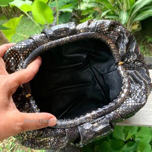 K&ouml;nnte beinhalten: Eine schwarze Clutch aus Pythonleder mit goldenen Akzenten und einem Verschluss. Die Clutch ist mit schwarzem Stoff gef&uuml;ttert.