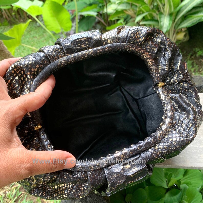 K&ouml;nnte beinhalten: Eine schwarze Clutch aus Pythonleder mit goldenen Akzenten und einem Verschluss. Die Clutch ist mit schwarzem Stoff gef&uuml;ttert.