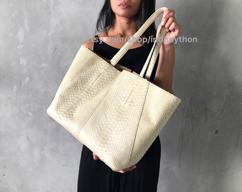 May include: A large off-white tote bag made of python skin. The bag has two top handles and a roomy interior.